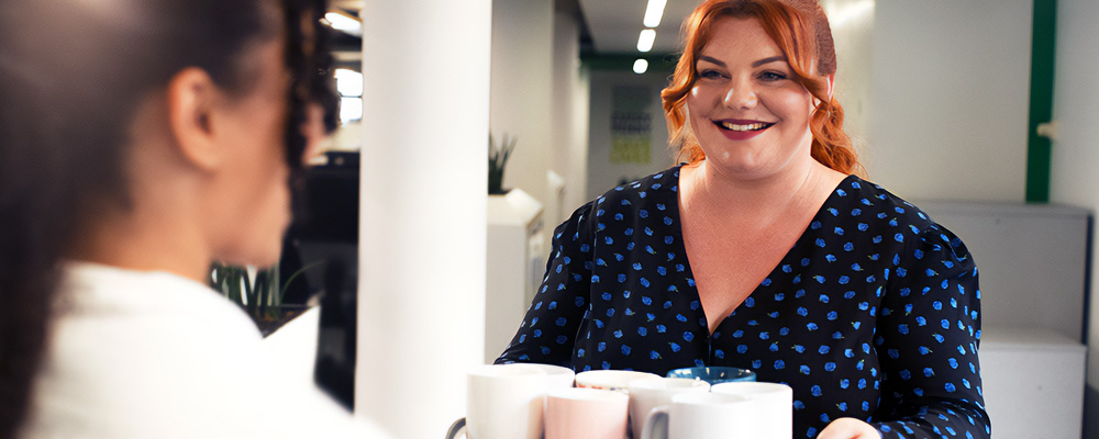 A woman carrying a tray with drinks and offering one to her colleague.